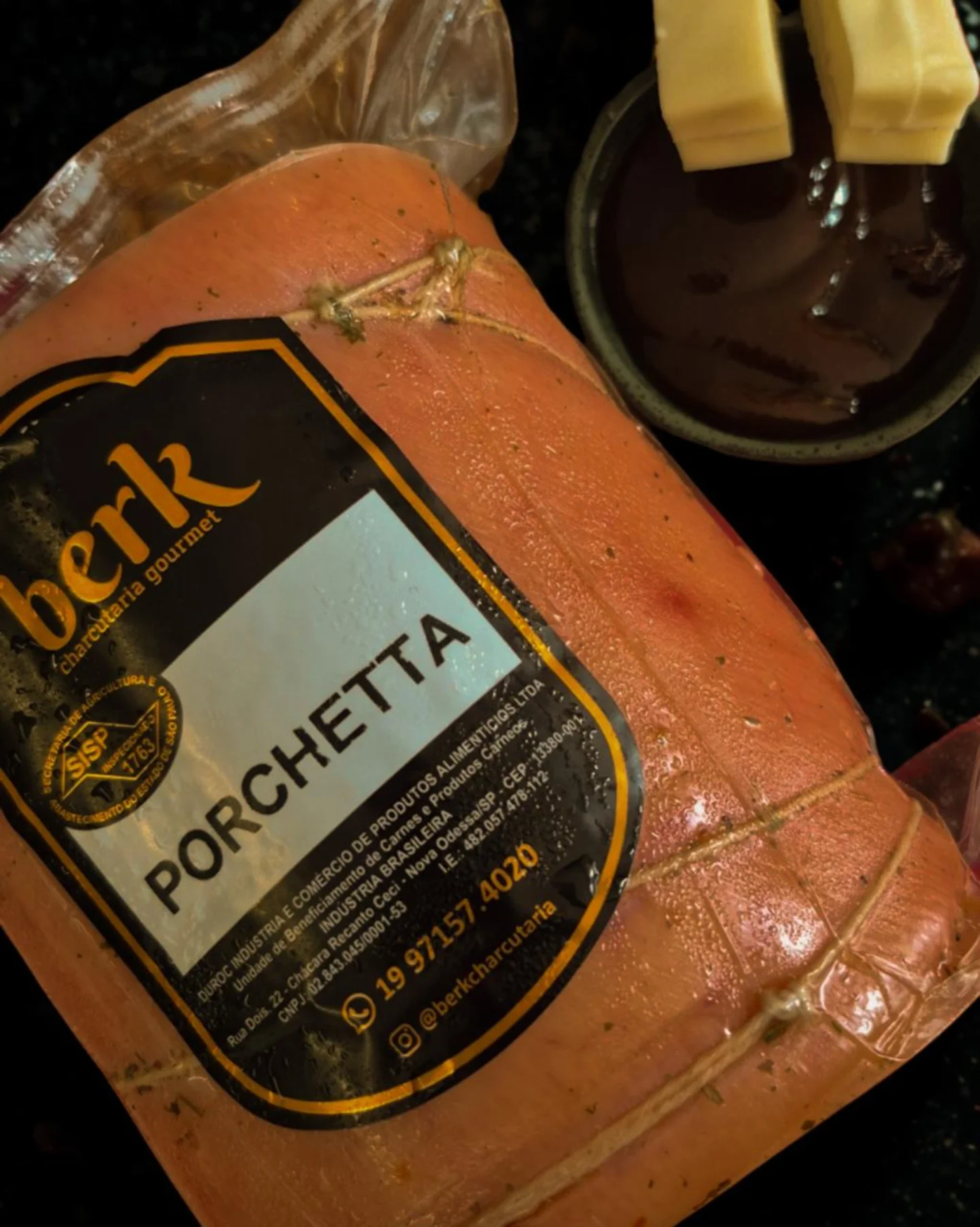 Peça de Porchetta Berk embalada a vácuo com rótulo, acompanhada de molho e queijo sobre fundo escuro